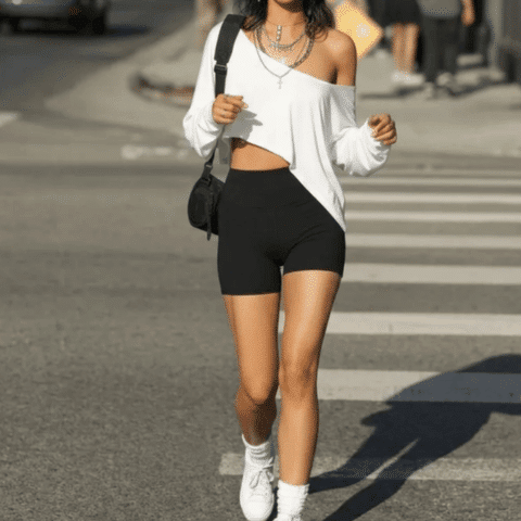 Tate McRae concert outfit idea: Woman wearing an off-shoulder white top, black biker shorts, white sneakers, and layered necklaces while walking on a city street.