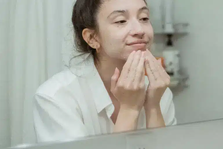 Woman applying facial cleanser in a bathroom with white shirt and hair tied up