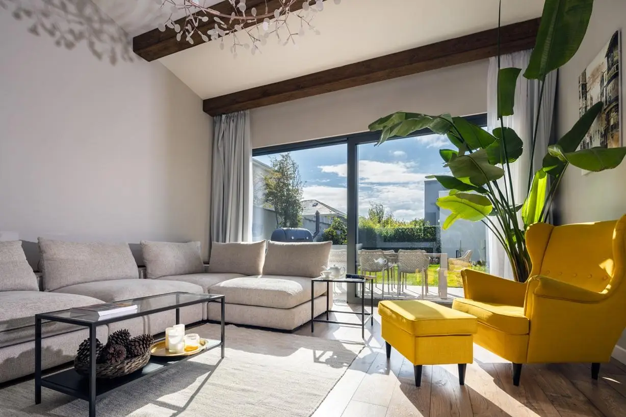 Modern living room with beige sectional sofa, yellow armchair, large plant, and glass doors to garden
