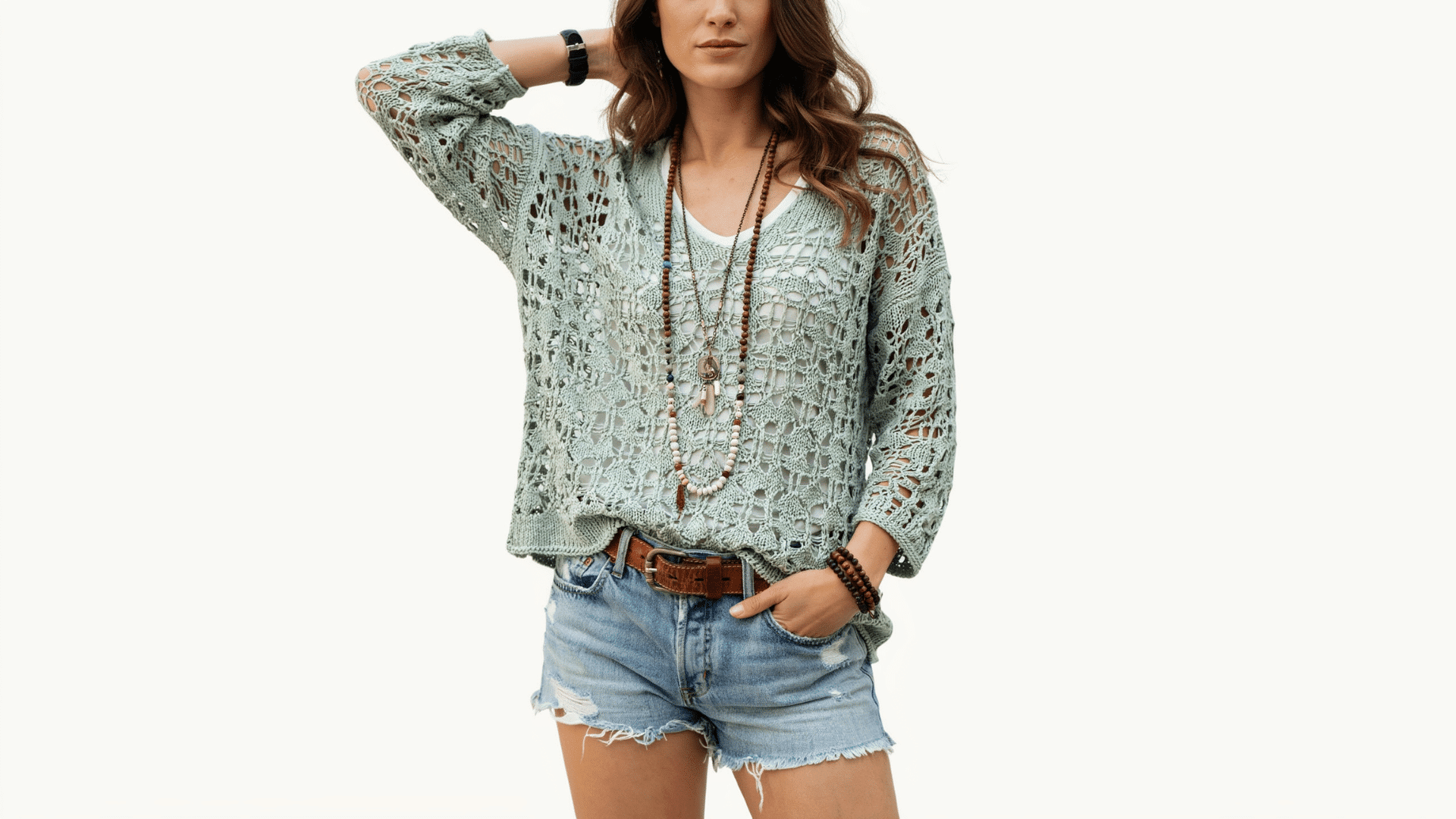 Woman in a boho sage green open-knit crochet sweater, denim shorts, and layered beaded necklaces on a white backdrop