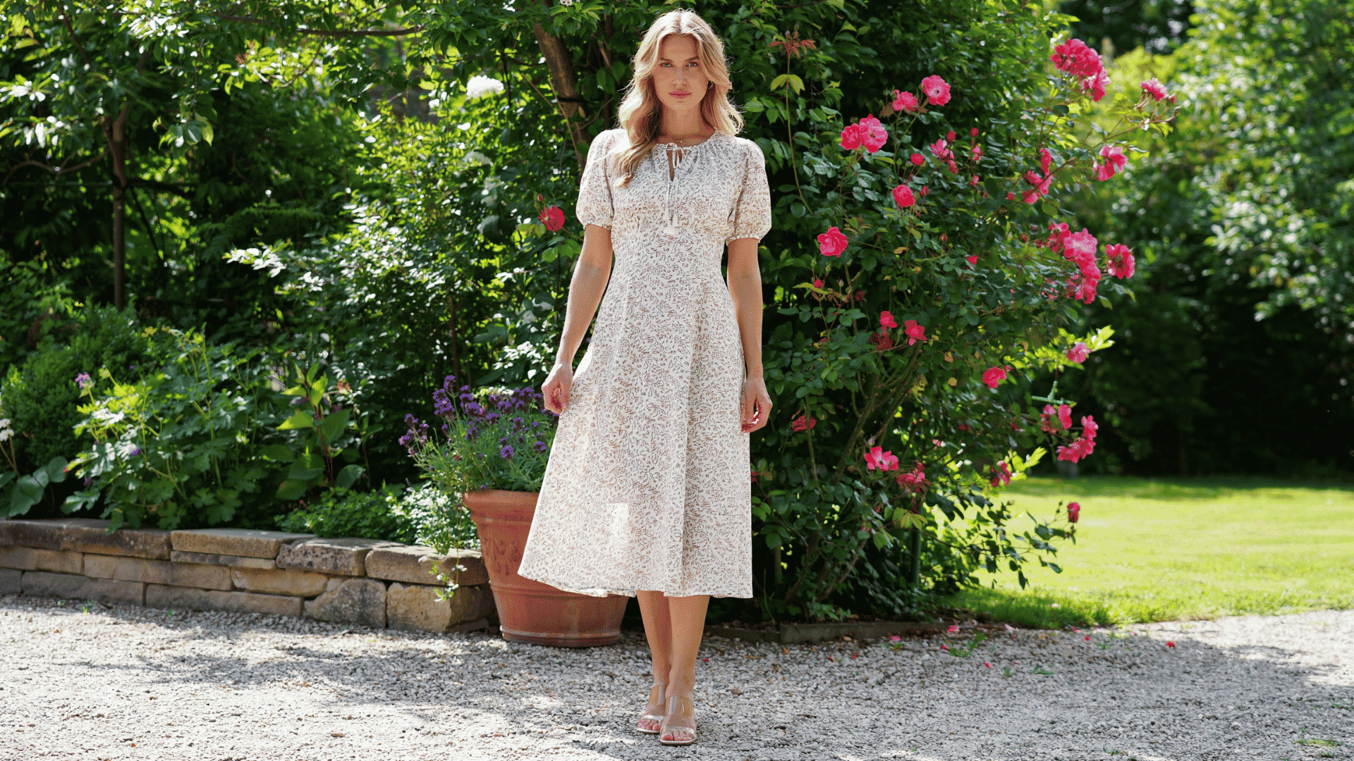 Woman in a boho style floral maxi dress with puff sleeves standing in a lush garden next to a pink rose bush