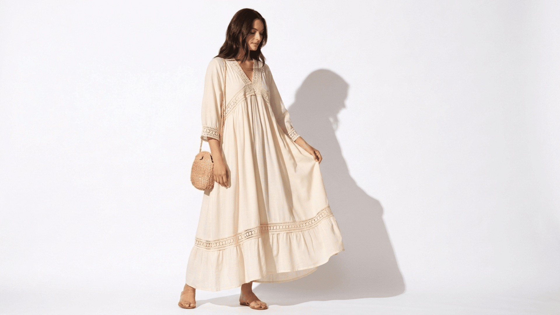 Woman in a cream linen boho maxi dress with lace inserts, sandals, and a straw bag against a white studio background