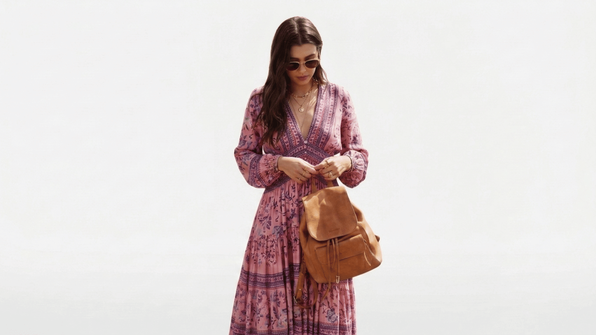 Woman in a pink floral minimalist boho maxi dress and sunglasses holding a tan leather backpack on a white backdrop