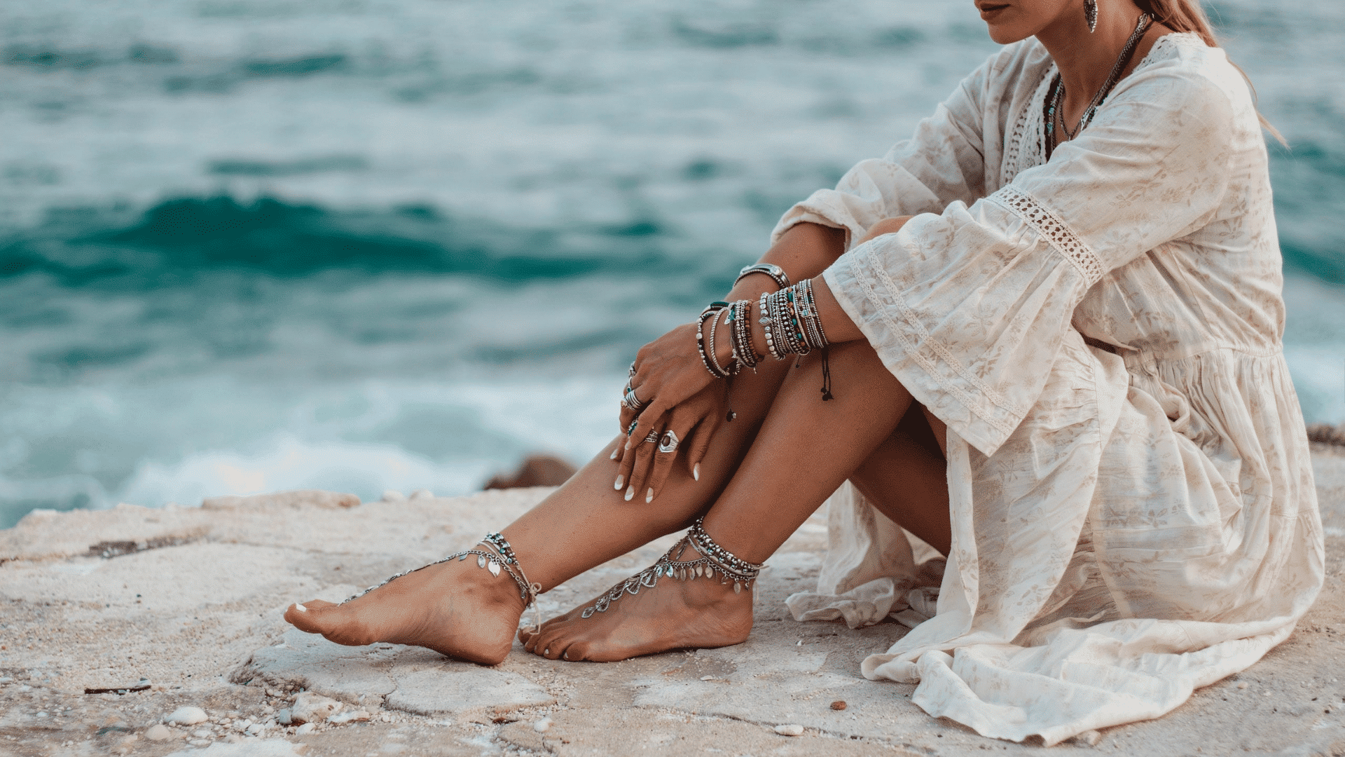 Woman in a white beach boho dress wearing stacked silver bracelets and ornate barefoot sandals by the ocean waves