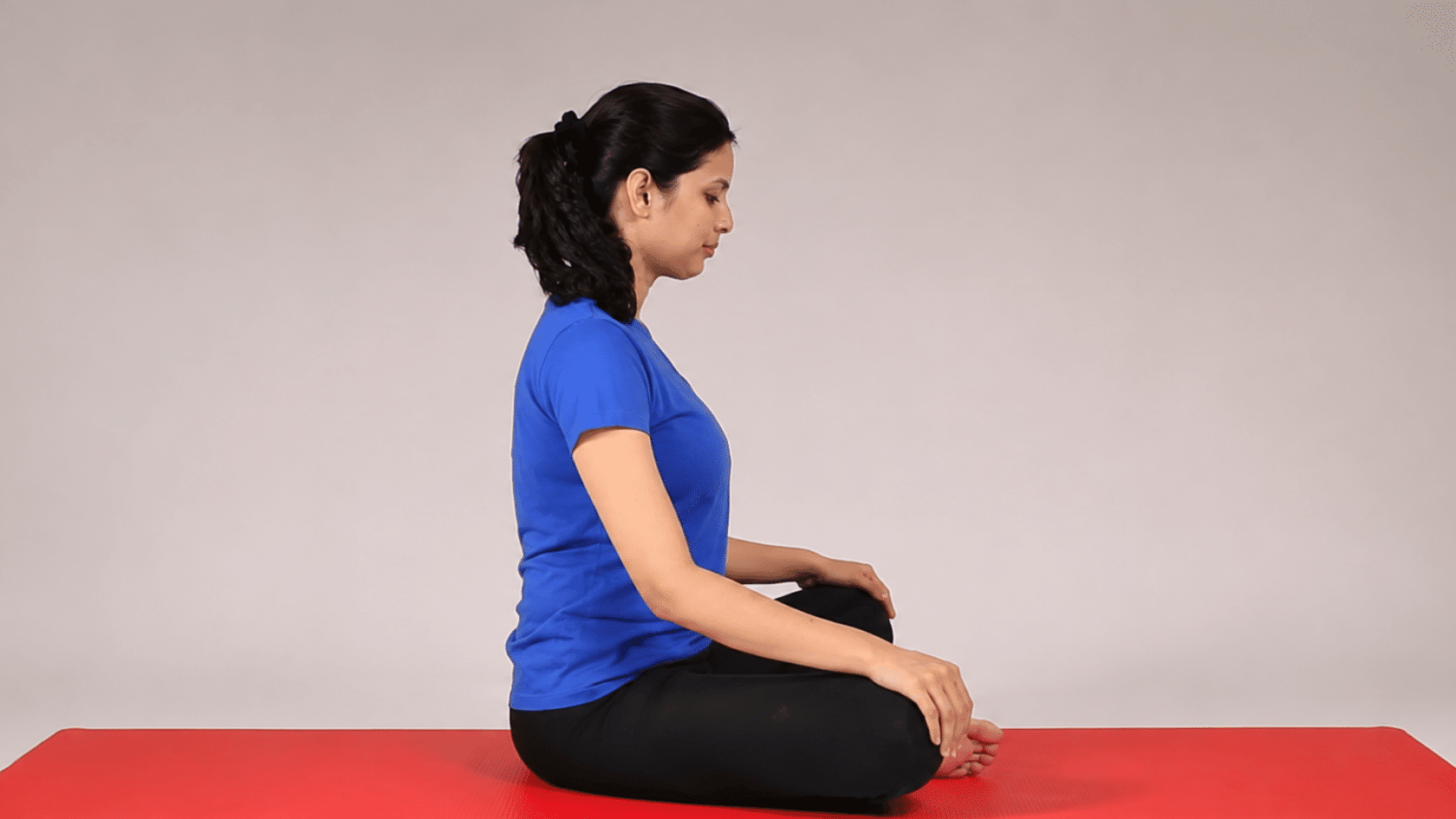 Woman sitting cross-legged on a red yoga mat in a studio setting.