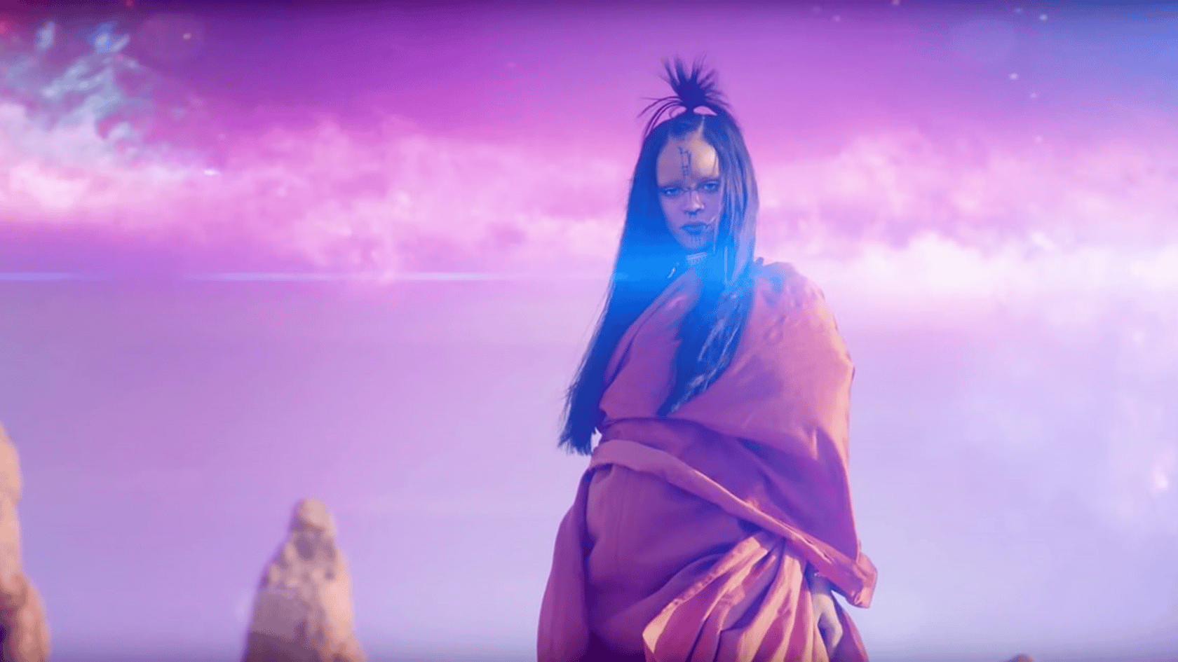 Woman standing in a flowing robe against a glowing purple sky with surreal, cinematic lighting and distant rock formations.