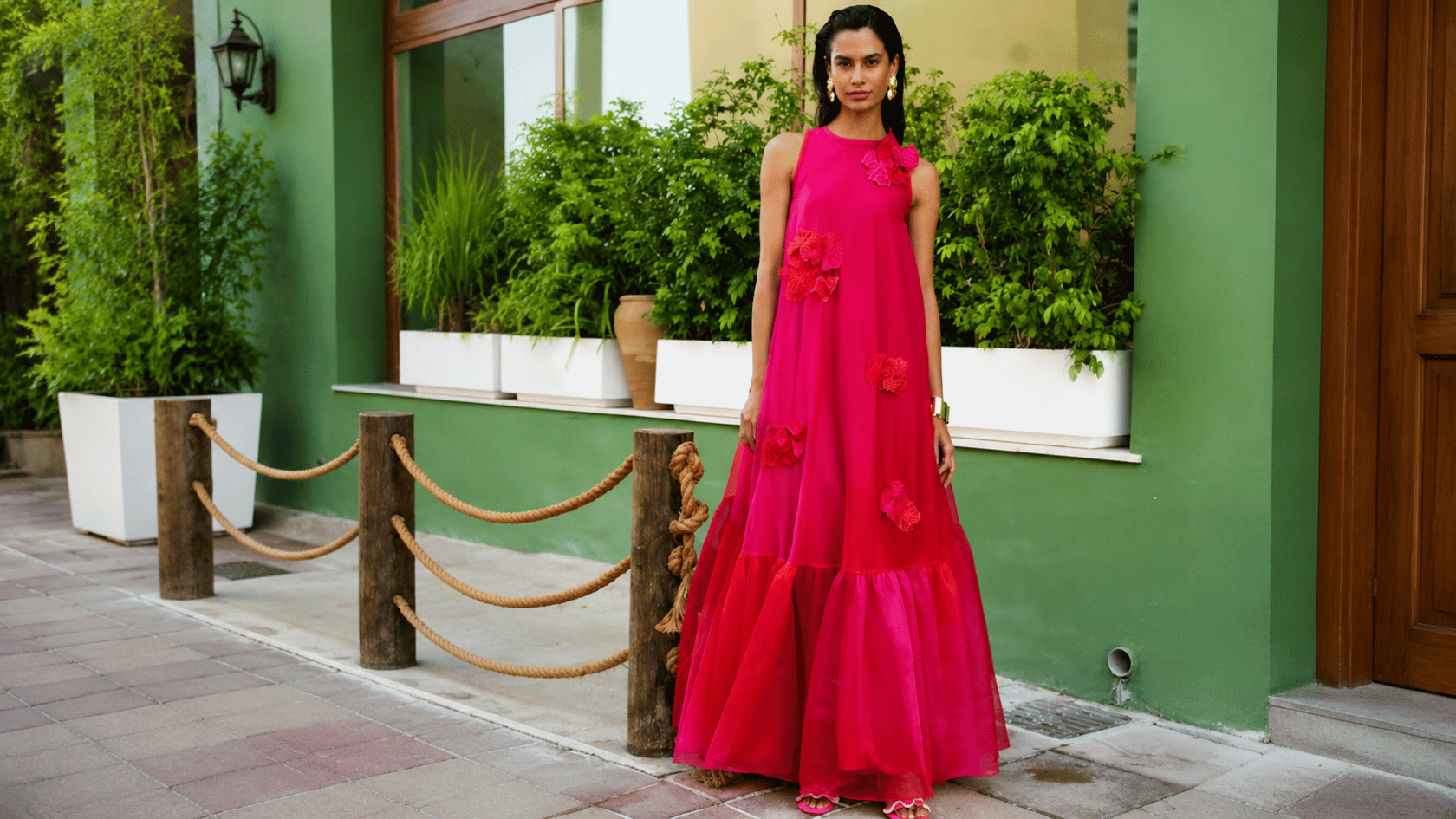 Woman wearing a pink A-line dress with floral details and a flared skirt standing outdoors near green walls and plants.