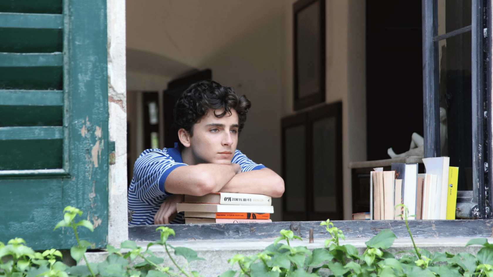 Young man resting his arms on stacked books at a window, looking outside thoughtfully
