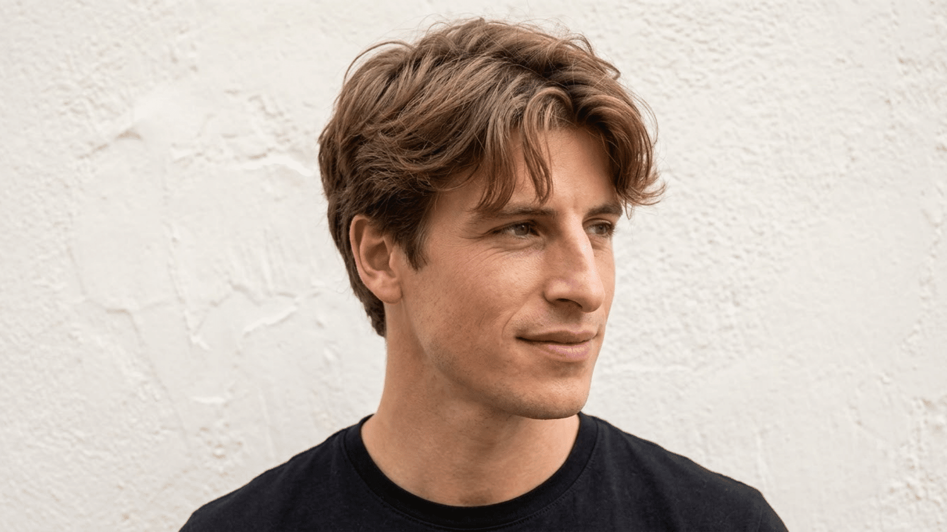 Young man with a textured curtain haircut wearing a black shirt against a light textured wall.