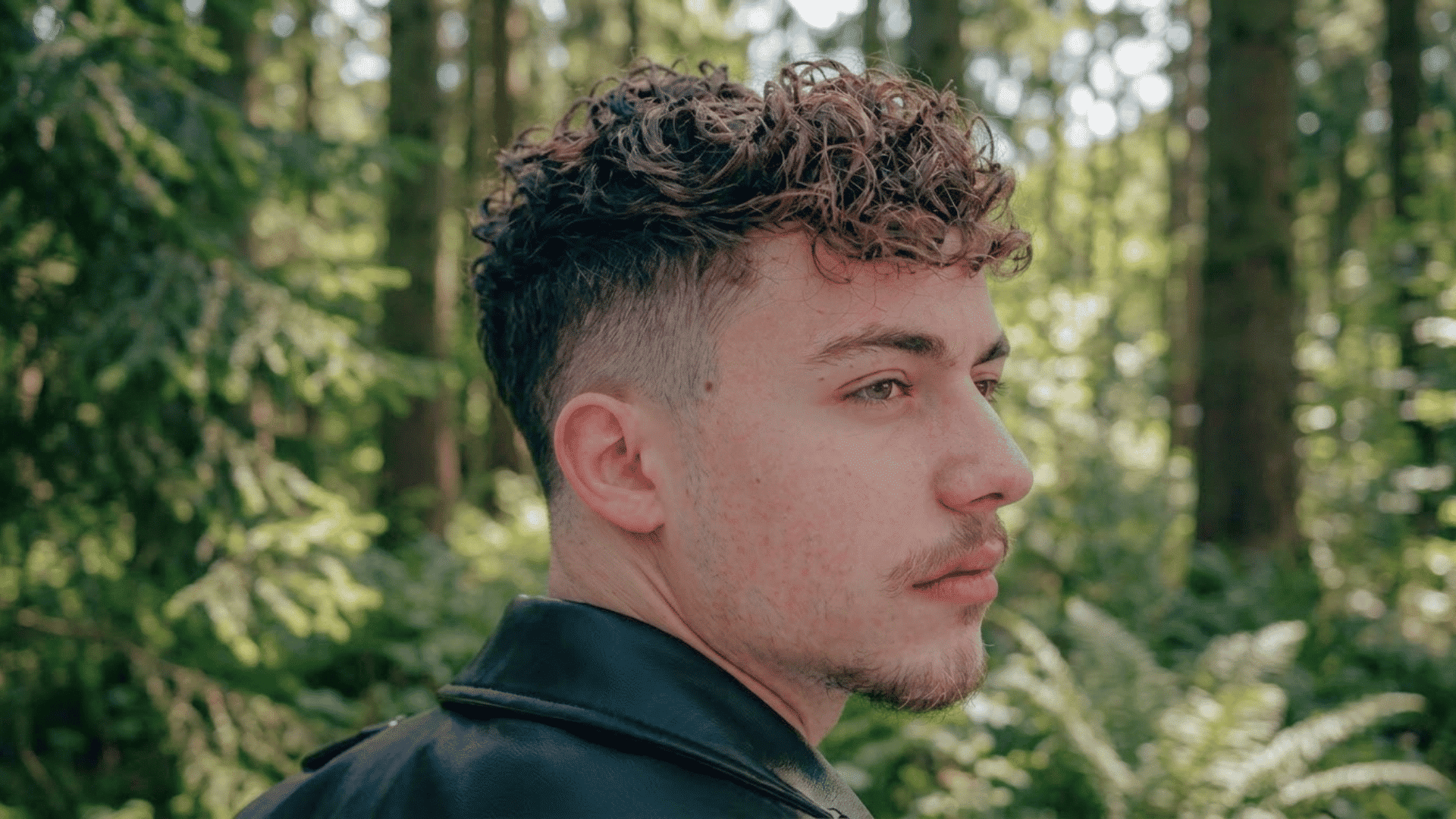 Young man with a wavy taper fade haircut viewed from the side in a forest setting.