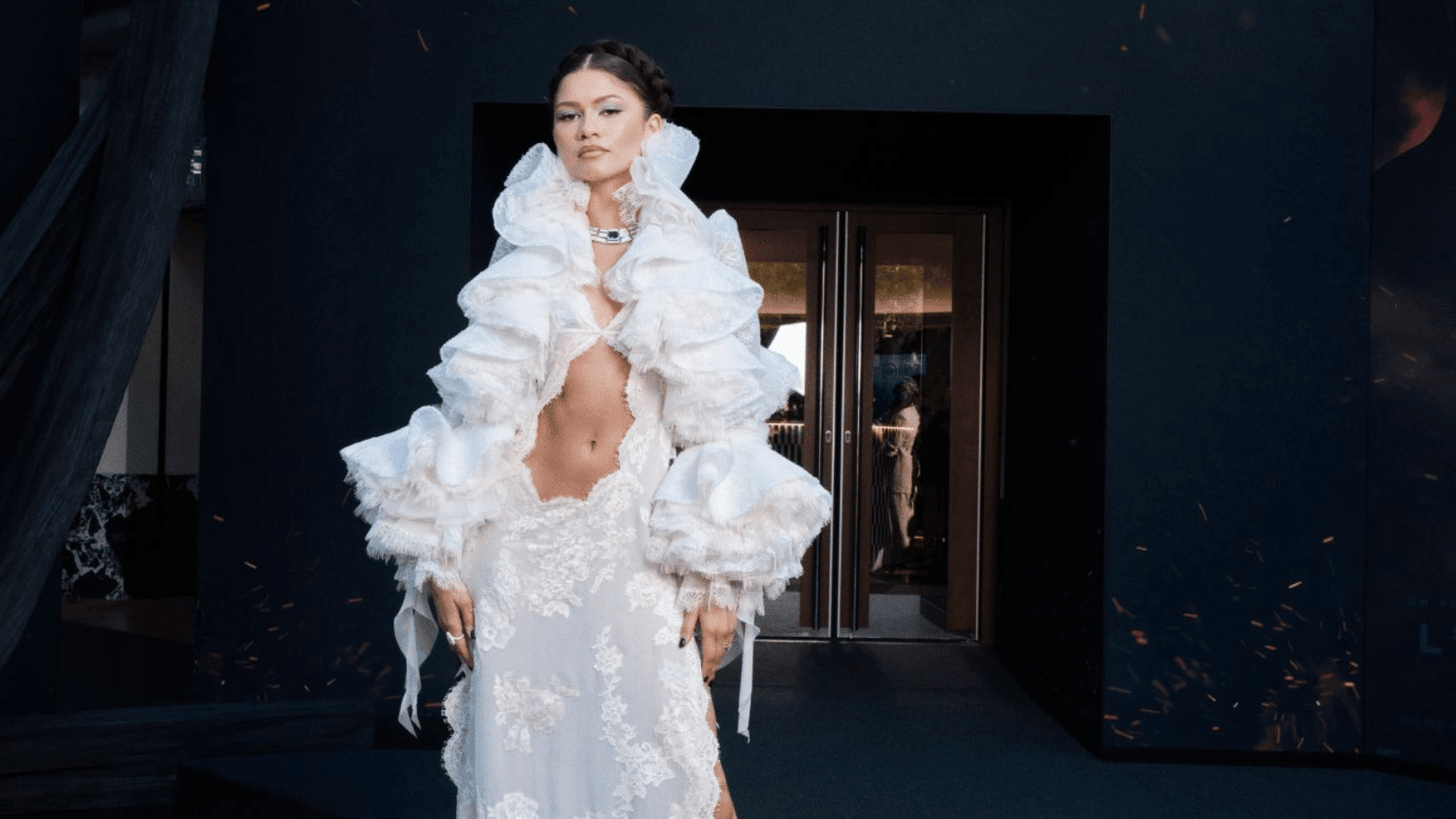 Zendaya wearing a white ruffled gown with a lace skirt at an indoor fashion event.