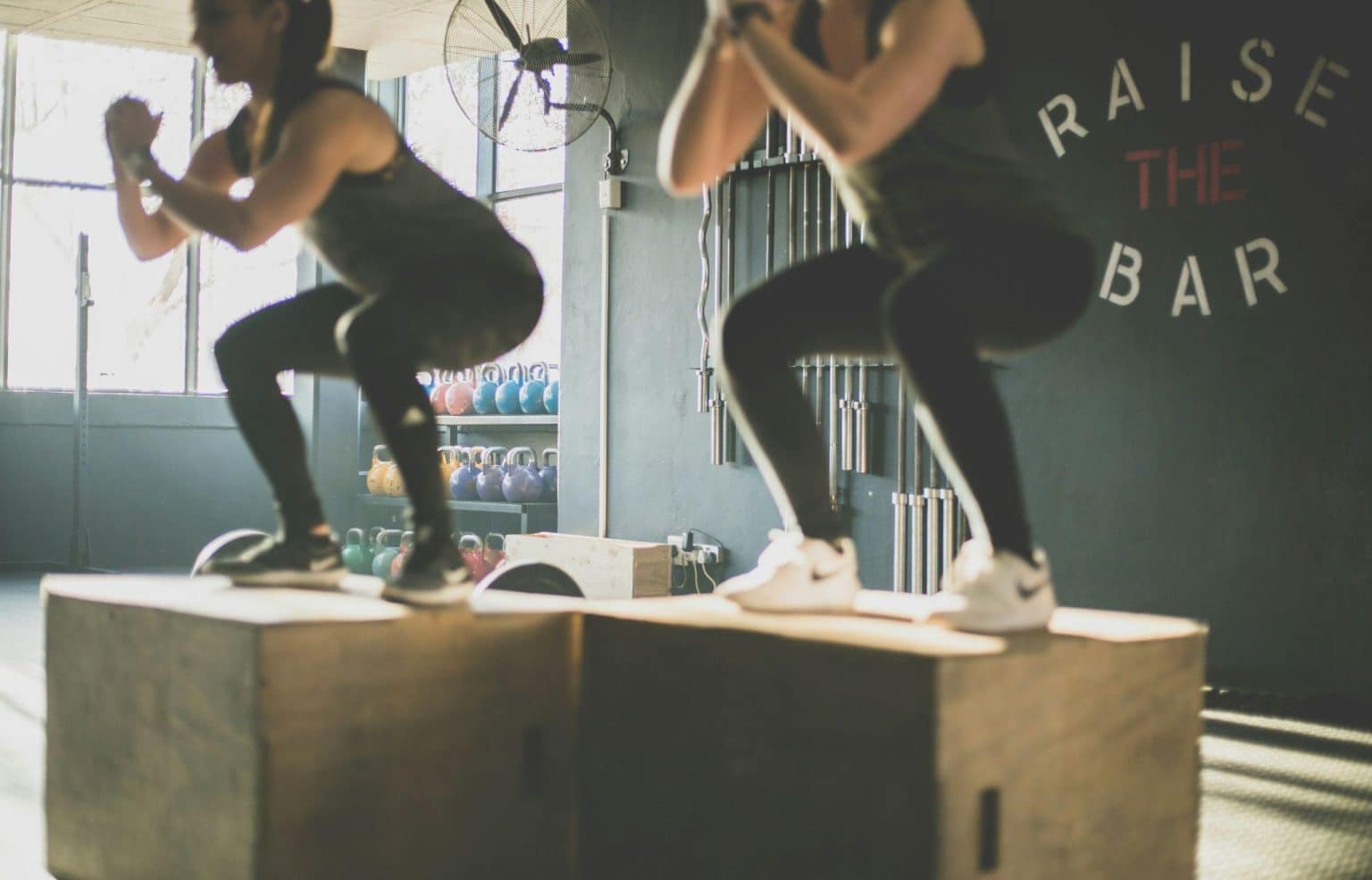 Two people performing box jumps in a gym with kettlebells and fitness equipment