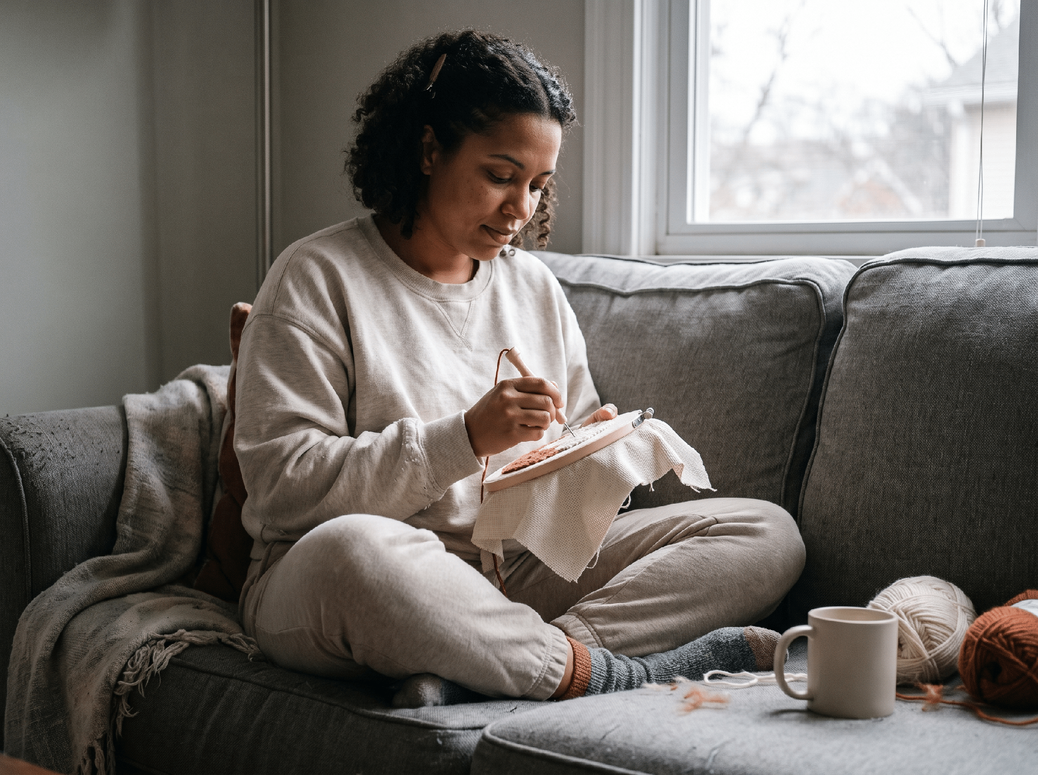 A person relaxed on a couch, working on a punch needle hoop in a cozy reading nook