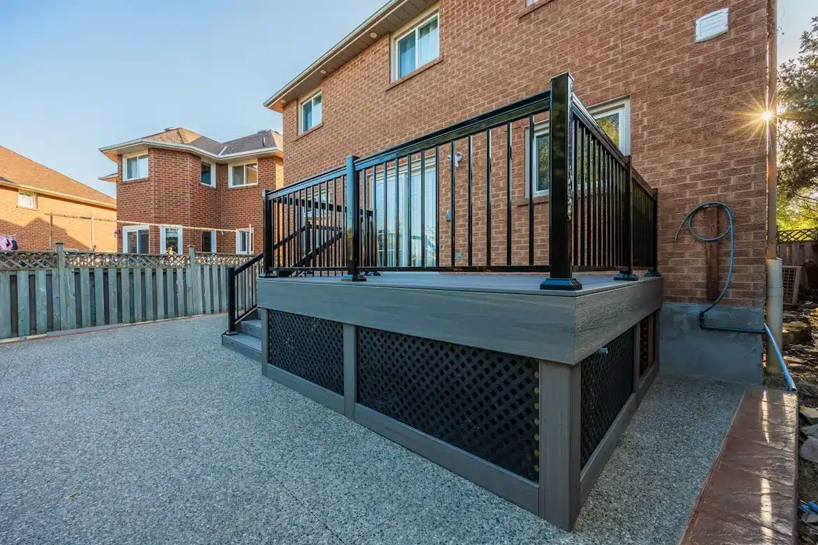 Elevated patio deck with black railing attached to brick house in sunny backyard setting