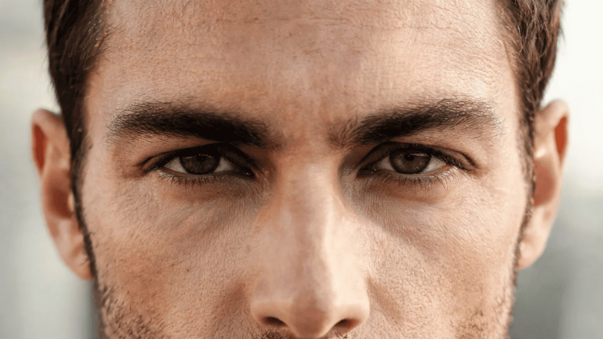 Close-up of a man with thick eyebrows, brown eyes, short facial hair, and ears visible