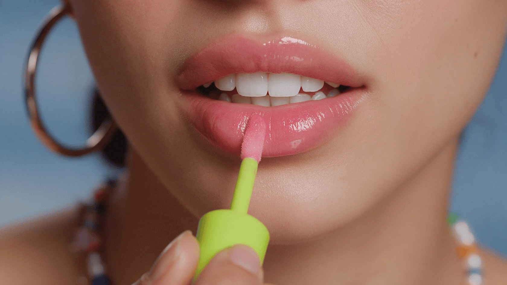 Close-up of a woman applying glossy pink lip oil to her lower lip with a wand applicator.