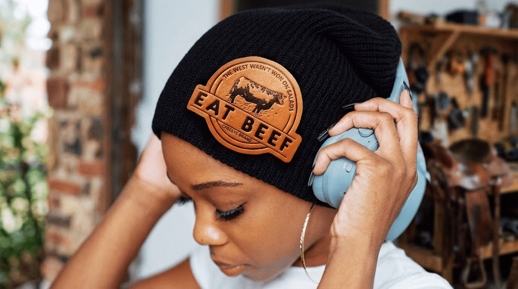 Black beanie with leather patch worn indoors, person adjusting blue headphones