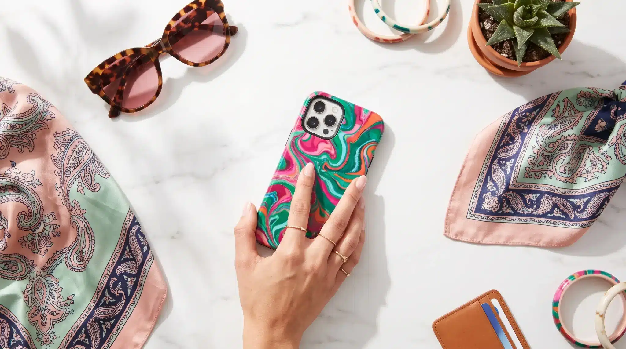 Hand holding phone with colorful case on marble surface surrounded by sunglasses and patterned fabric