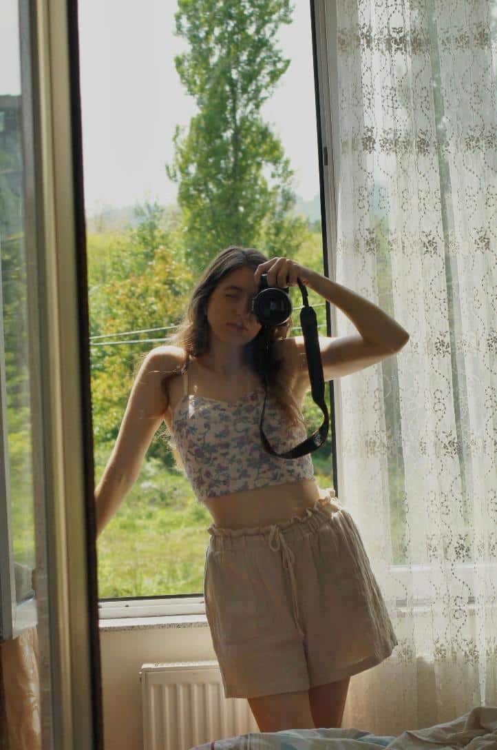 Person taking mirror selfie in sunlit room with floral top and beige shorts