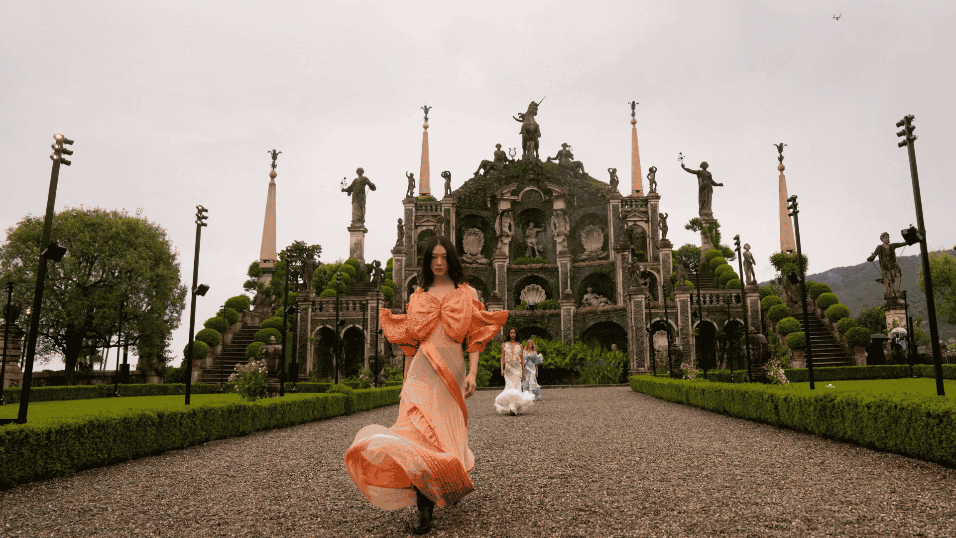 Model in orange flowing dress walking on gravel runway in garden with ornate baroque architecture and statues behind