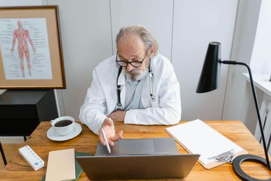 Doctor with stethoscope using laptop at desk in medical office with anatomy chart