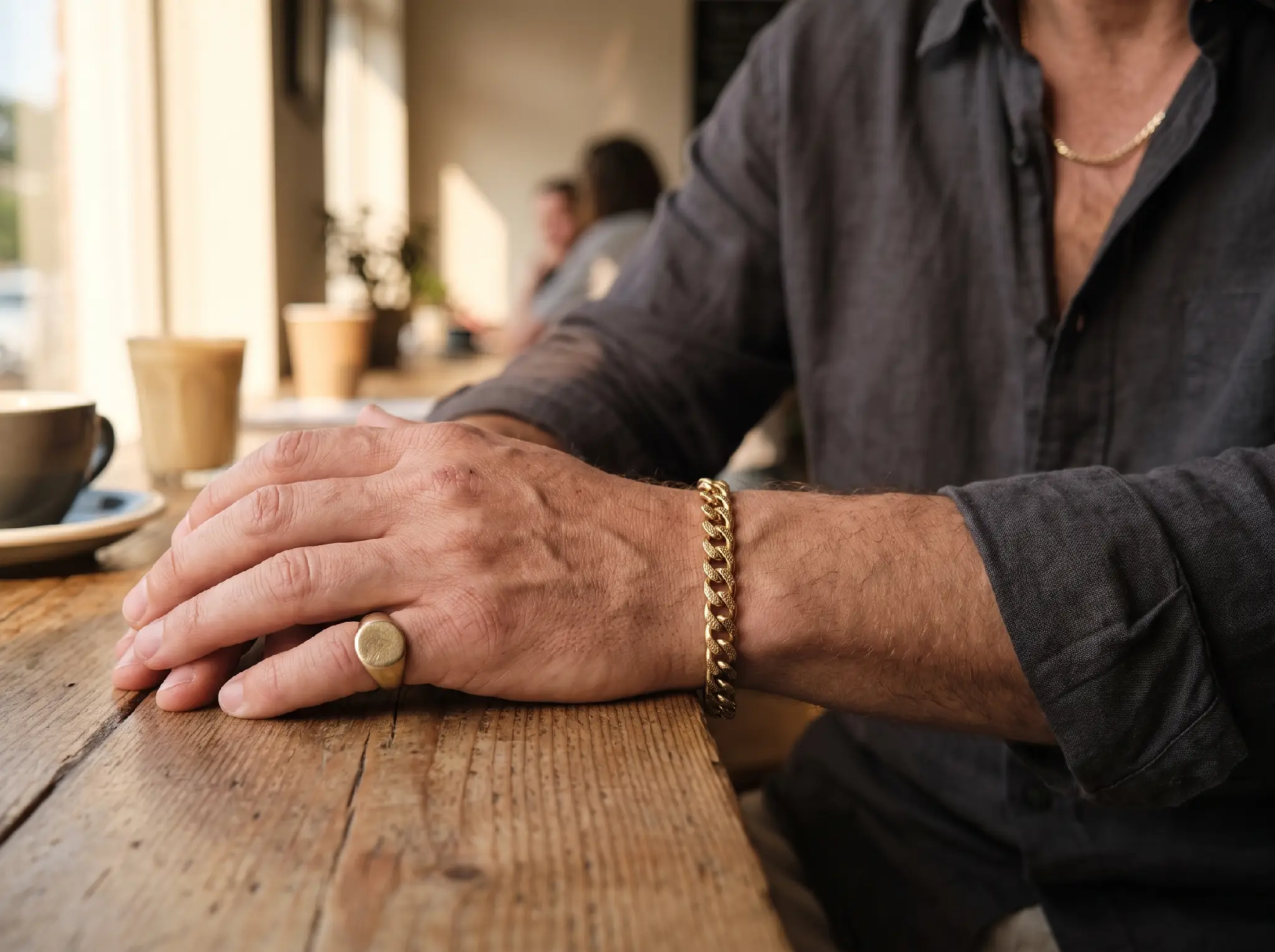Close-up of a man's hands with gold jewelry resting on a wooden table in a café setting