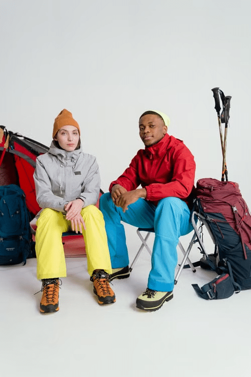 Two people in winter gear seated indoors with hiking backpacks and trekking poles nearby