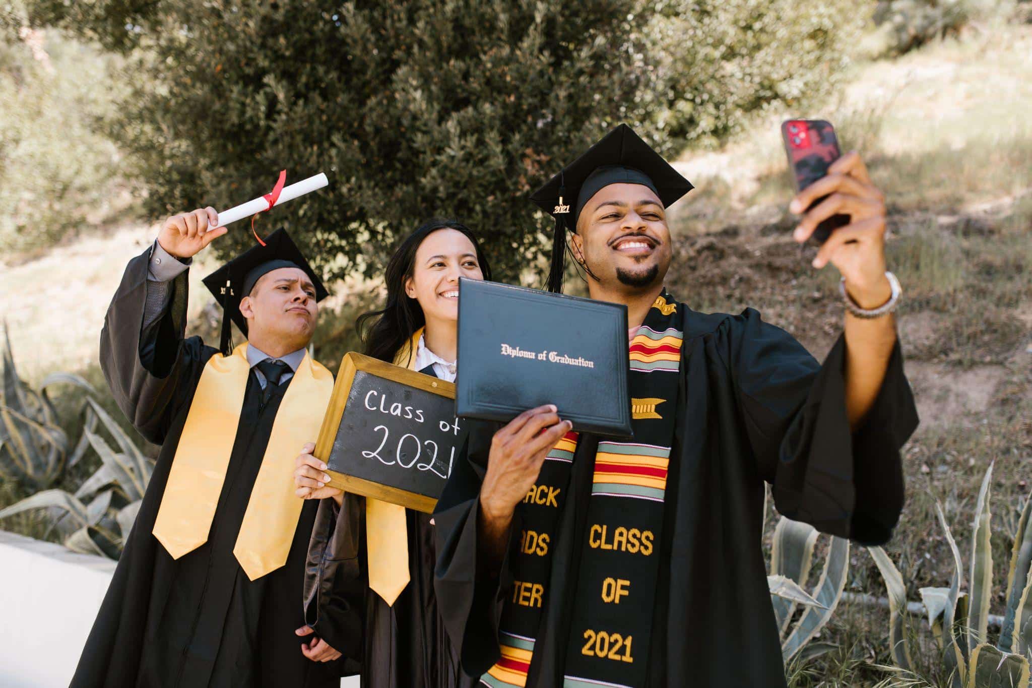 Graduates in caps and gowns taking a selfie outdoors with diplomas and chalkboard sign