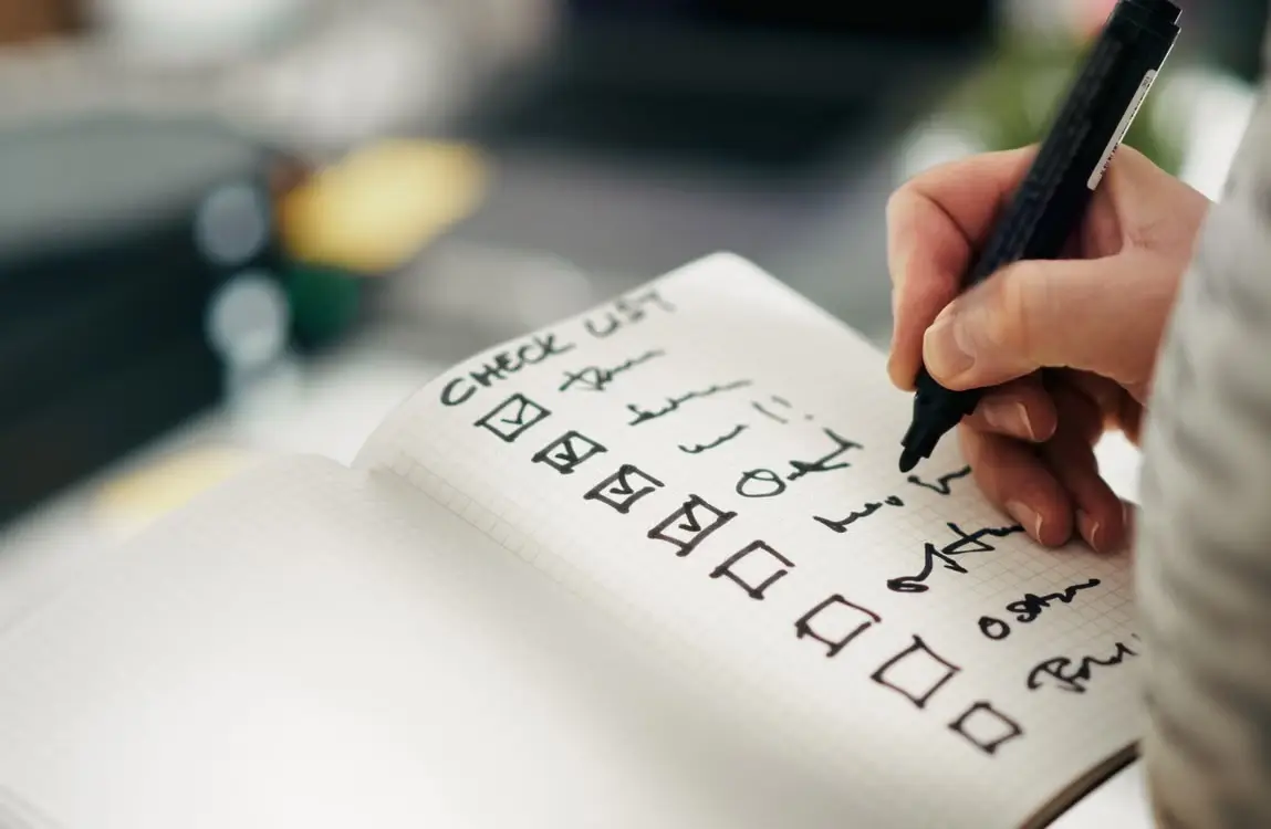 Hand writing checklist in notebook with black marker in blurred office setting