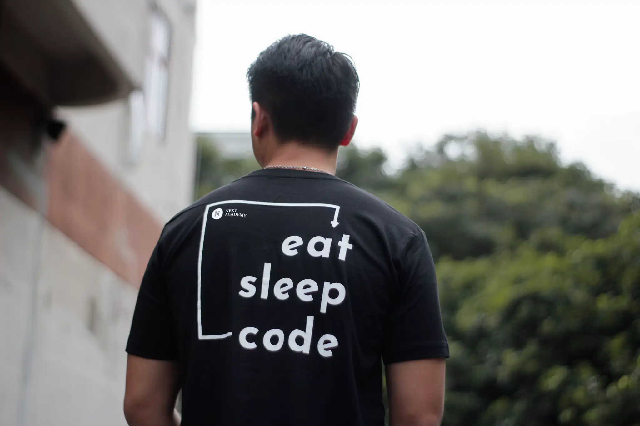 Man wearing black t-shirt with "eat sleep code" text standing outdoors near trees