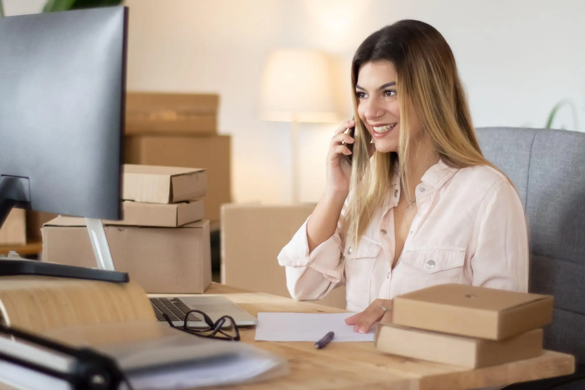 Smiling woman on phone at desk with cardboard boxes and computer monitor