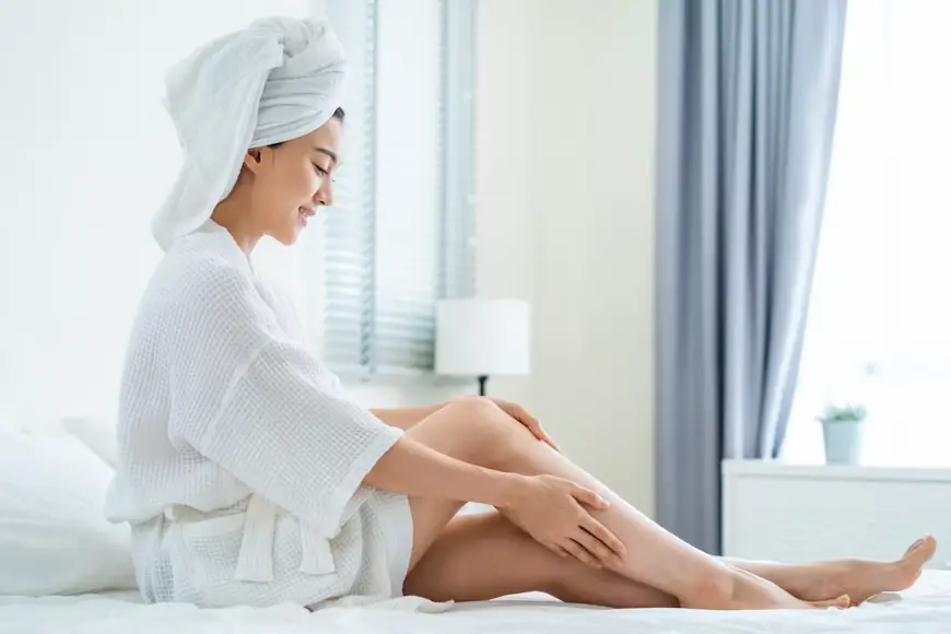 Woman in white robe with towel on head sitting on bed in bright bedroom