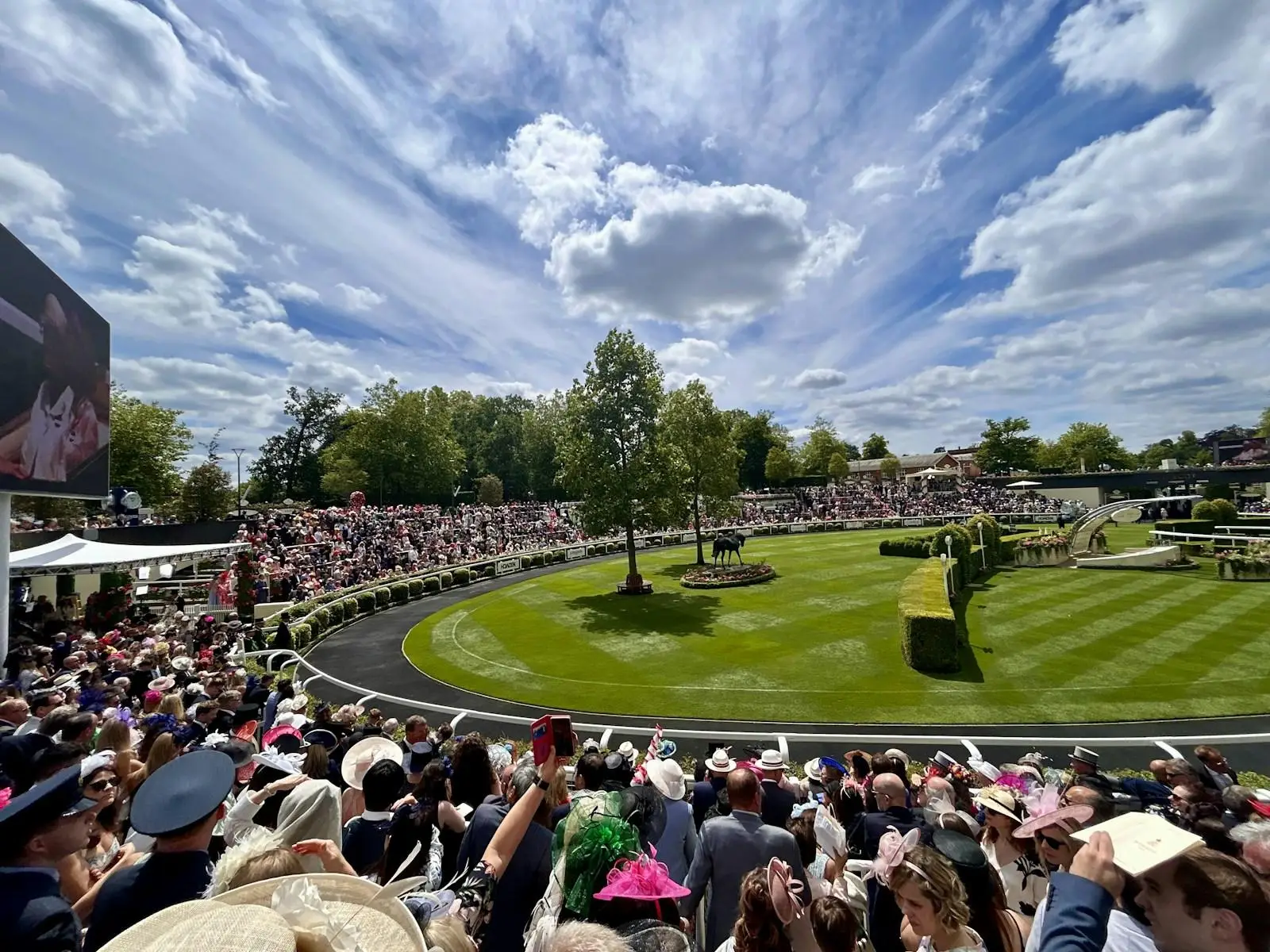 Crowd in hats watching horse track with manicured lawn and cloudy sky overhead