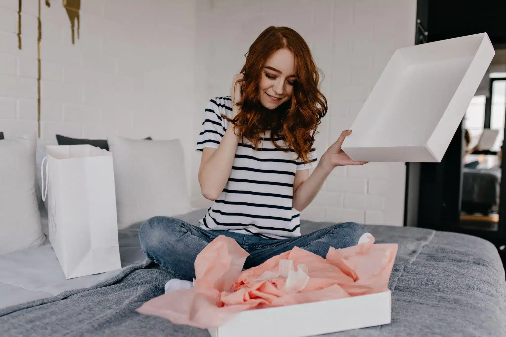 Woman opening white gift box with pink tissue paper on bed in cozy bedroom