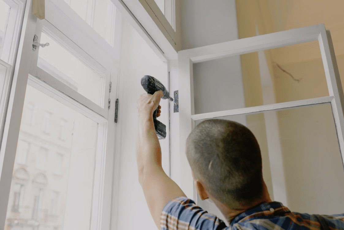 Man in plaid shirt using power drill to fix window frame indoors